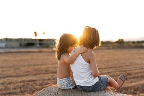 Two children sit together on a hay bale in the warm glow of a sunset, embracing and enjoying the