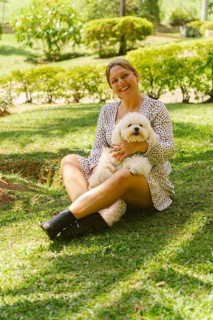 Happy adult woman sitting on lush green grass in a park, joyfully embracing her small white poodle
