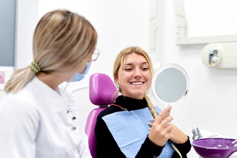 A confident woman smiles while holding a mirror during a dental check-up. A friendly dental