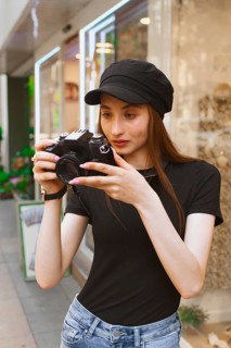 A young woman in casual attire, wearing a black cap, focuses intently on her camera as she takes