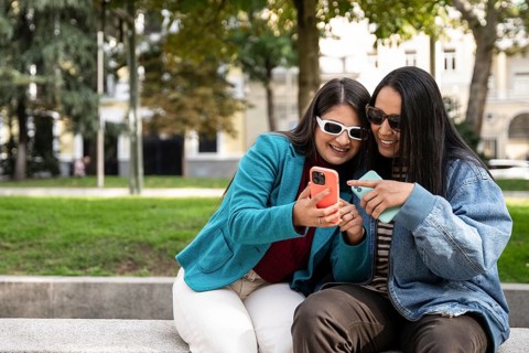 A joyful Latin lesbian couple sits on a park bench, sharing a smartphone. They wear casual outfits