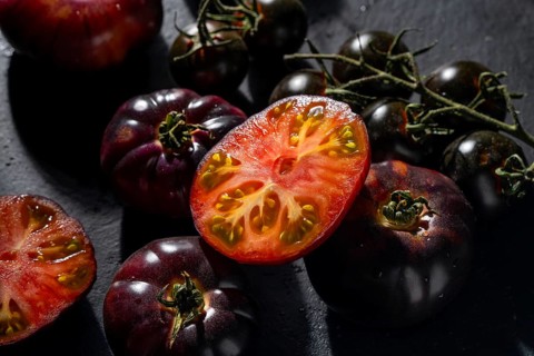 Fresh heirloom tomatoes are displayed on a dark surface. The vibrant red and purple skins contrast