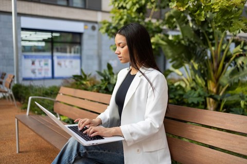 A professional woman in a white blazer works on a laptop, seated on a wooden bench in an urban park