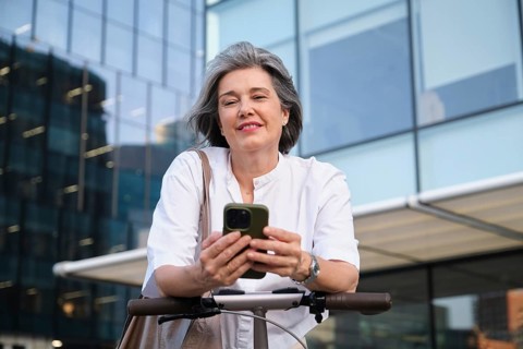 Mature woman in a stylish white blouse smiles while using a smartphone outdoors, leaning on an