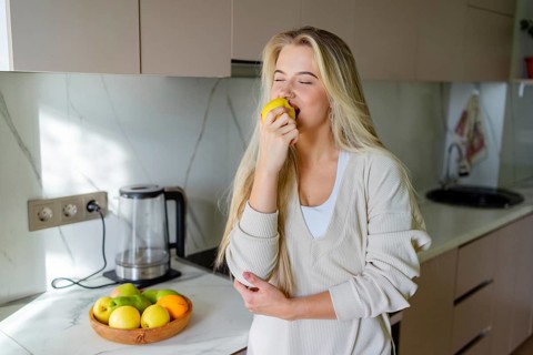 A woman stands in a stylish kitchen, savoring a juicy apple. Beside her is a bowl with various