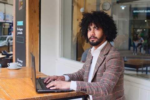 Mixed-race insurance salesman with afro hair using a laptop at a wooden table in a café. Dressed in