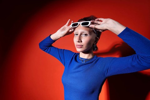 A stylish woman in a bright blue top poses confidently against a striking red background, adjusting