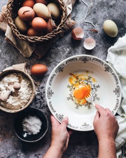 Rustic kitchen scene depicting a person preparing to bake. A cracked egg is being whisked in a