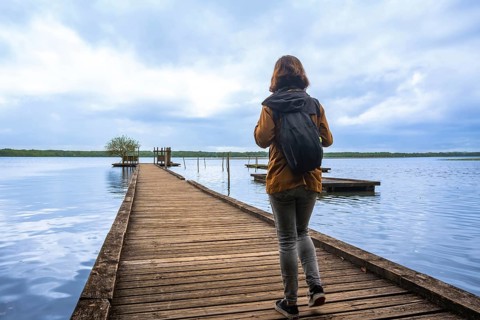 Woman backpacker walking on wooden pier into calm blue lake azur under cloudy sky, evoking travel,