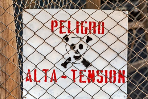 Spanish warning sign with skull and lettering 'Peligro Alta Tension' behind a chain link fence,