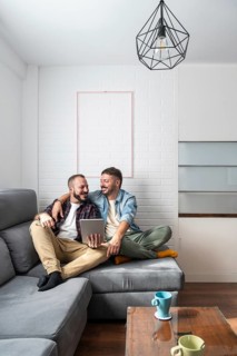A joyful gay couple sits together on a comfortable couch in their modern living room, using a