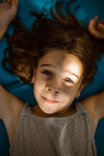 A child lies on a blue surface, smiling softly as sunlight casts delicate shadows on their face.
