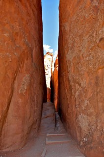 A narrow passage of a hiking trail between high sandstone walls, into which light is just falling,