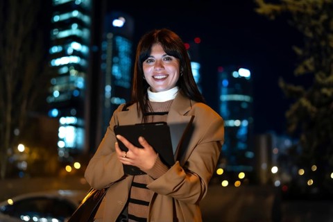 Young professional businesswoman smiling, holding a digital tablet outdoors at night in a city