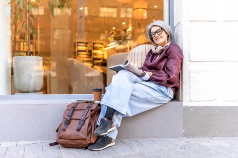 Active mature woman with gray hair wearing glasses and headphones, sitting on a window ledge,