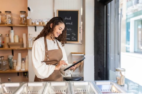 A cheerful woman clerk wearing a brown apron uses a tablet to manage orders in a modern ice cream