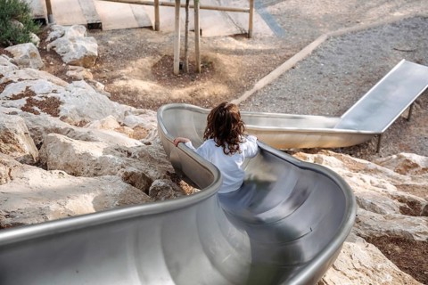 A young girl in a white shirt slides down a metal slide in a forest park on a sunny afternoon,