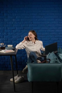 A woman in a white shirt and jeans sits on a couch, multitasking with a laptop and phone,