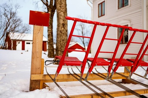 Vibrant red traditional kick sleds are lined up against a wooden post, set in a snowy landscape.