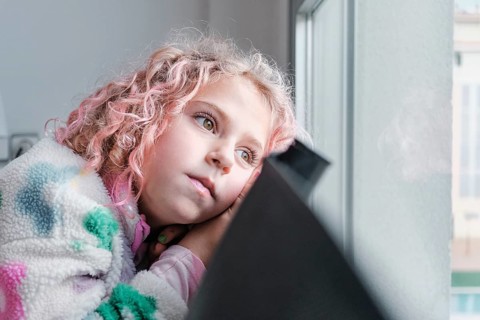 A young child with curly pink hair looks thoughtfully out a window. Wrapped in a cozy, colorful