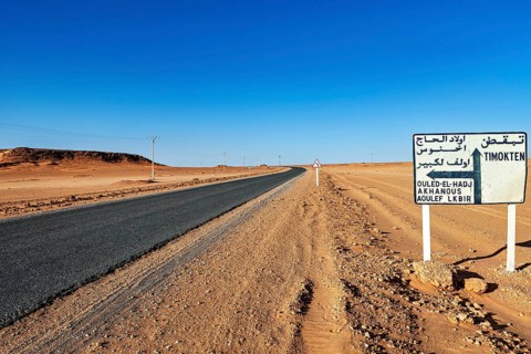 Road leads through a barren desert landscape under a clear blue sky with a signpost in Arabic, By