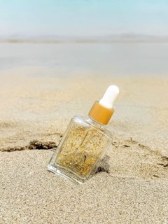 A hair care product with gold flecks sits embedded in the sand against a serene sea backdrop on a