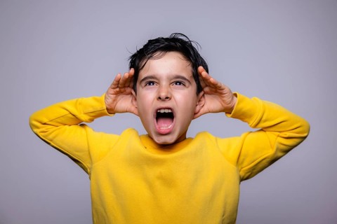 A young boy in a yellow sweater energetically shouts with hands raised to his ears, expressing a