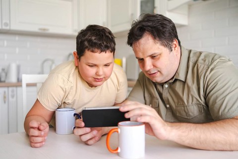 A father and son sit at a kitchen table, both focused on a smartphone. They share quality time,