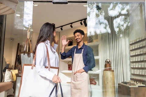A customer exchanges a friendly greeting with a salesperson in a trendy bag store. Shelves showcase