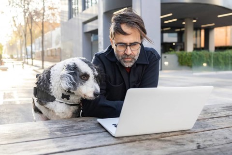 A man sits at a wooden table in an urban setting, focused on his laptop with his pet dog beside him