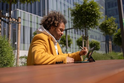 A man with afro hair in a yellow coat uses a tablet outdoors. Seated at a wooden table, he appears