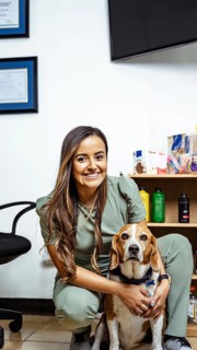 A nutritionist in a green uniform poses with her dog in a professional office setting. Certificates