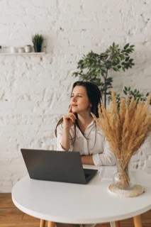 A nutritionist sits thoughtfully at a white desk with a laptop and pampas grass in a vase. The
