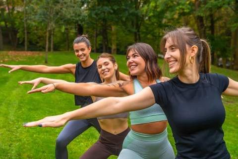 Group of smiling women practicing yoga together in a green park, doing warrior poses and stretches