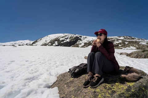 An Asian woman sits on a rock in the snow-covered Sierra de Gredos, Spain, enjoying a snack under