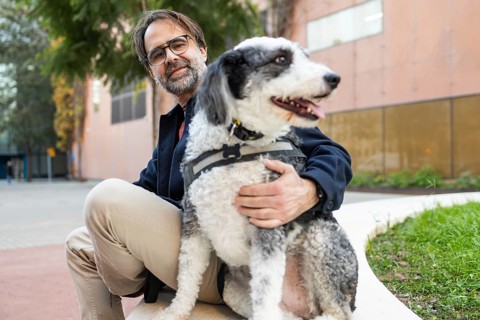 A cheerful man with a beard is seen bonding with his black and white dog while sitting in a city