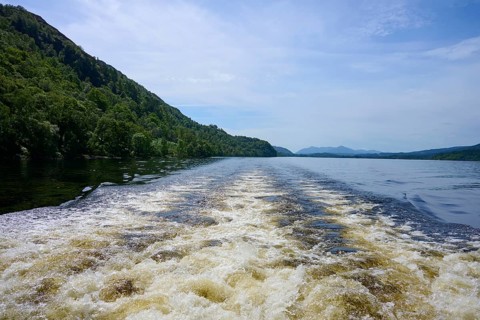 A river with waves from a boat surrounded by green hills and a blue sky, Fort Augustus, Loch Ness,
