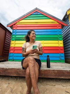 A woman with a smartphone sits relaxed in front of vibrant Brighton Bathing Boxes in Melbourne,