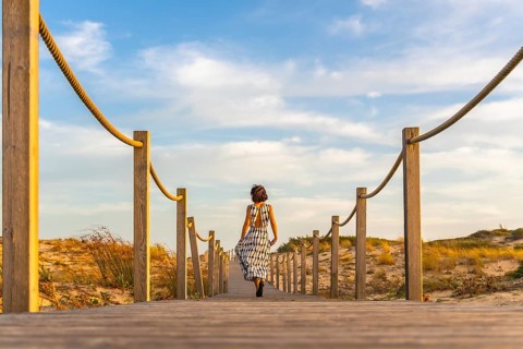 Woman walking away on a wooden boardwalk through sand dunes toward the atlantic beach at seignosse
