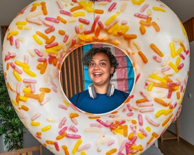 A joyful transgender person with a playful expression peeks through a large decorative donut float,