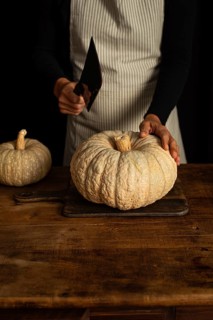 A person in an apron prepares to cut a large pumpkin on a wooden table. The warm, rustic setting