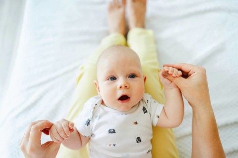 A baby in a patterned onesie lies on a mother lap, holding their hands. The setting is cozy and