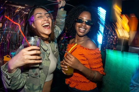 Two cheerful diverse women hold drinks and celebrate during a vibrant evening at a nightclub,