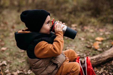 A child dressed warmly in a black beanie and brown outfit drinks from a thermos while sitting on