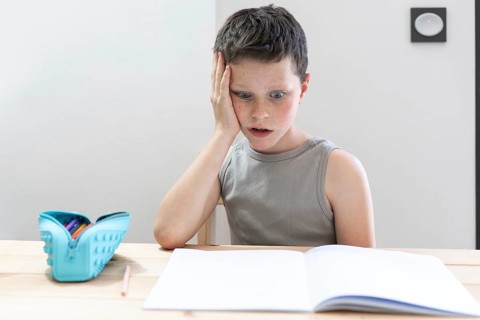 A young boy with a surprised expression sits at a desk with an open book, staring at the pages. A