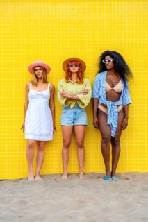 Three diverse young women wearing summer clothes and hats are posing on a sandy beach with a