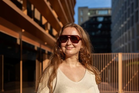 A woman with long hair and sunglasses enjoys the warm sunlight against an urban backdrop. Her