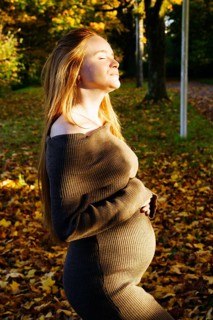 A beautiful pregnant woman with long red hair stands in a sun-dappled autumnal setting, her eyes