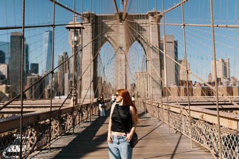 A woman with red hair walks across the Brooklyn Bridge, basking in the sun. The iconic bridge