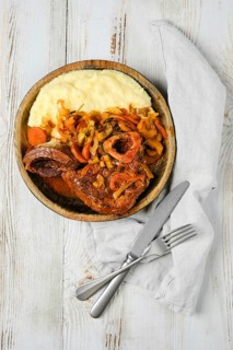 A wooden bowl filled with osso buco, mashed potatoes, and vegetables. A knife and fork are placed